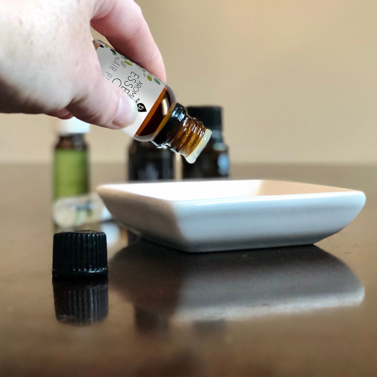 Close-up of a hand tilting an essential oil bottle over a white ceramic dish, with other aromatherapy oil bottles in the background.